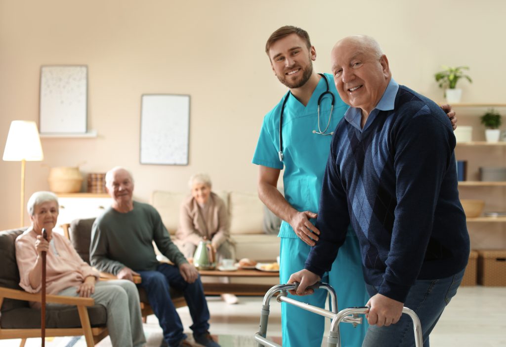 Care Worker Helping To Elderly Man With Walker In Geriatric Hosp